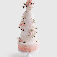A tall, elegant white tiered cake decorated with pink and white sugar flowers and green leaves, featuring intricate textures and floral arrangements, displayed on a white cake stand against a light background.