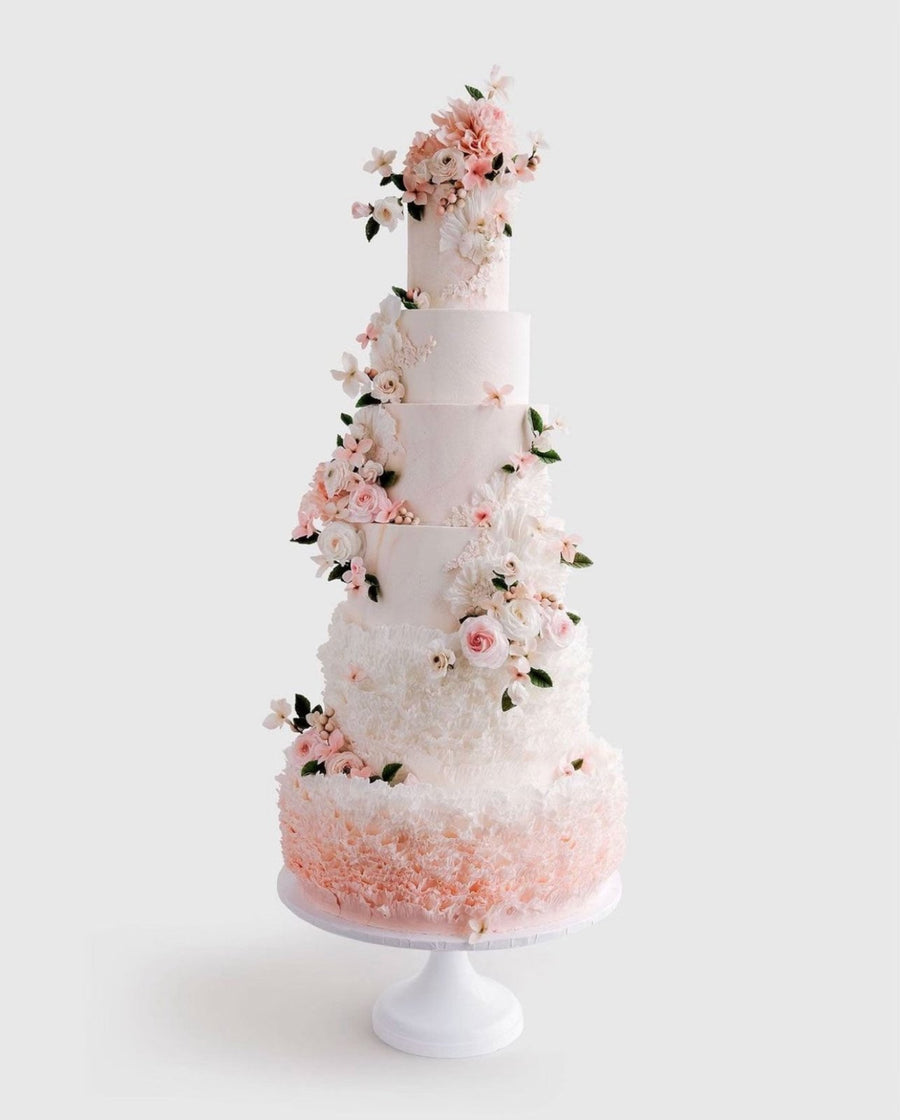 A tall, elegant white tiered cake decorated with pink and white sugar flowers and green leaves, featuring intricate textures and floral arrangements, displayed on a white cake stand against a light background.
