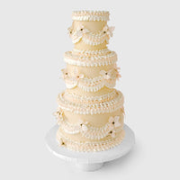 Jenna Rae Cakes’ Wedding Cakes Under $1,000 features a four-tier cream-colored cake with scalloped icing and delicate flower decorations, presented on a white stand against a light backdrop.