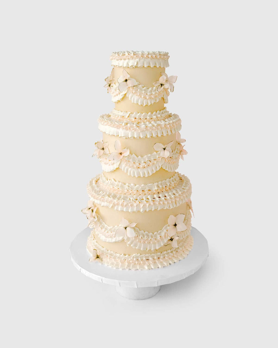 Jenna Rae Cakes’ Wedding Cakes Under $1,000 features a four-tier cream-colored cake with scalloped icing and delicate flower decorations, presented on a white stand against a light backdrop.