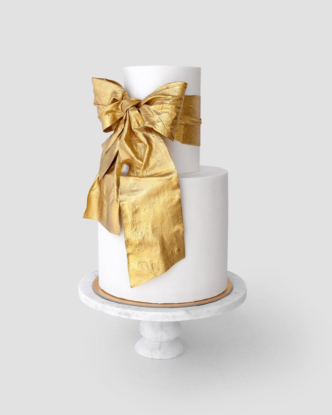 Jenna Rae Cakes’ “Wedding Cakes Under $1,000”: A two-tier white cake on a marble stand, topped with a statement metallic gold bow.