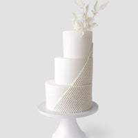 The Wedding Cakes Under $1,000 by Jenna Rae Cakes features a three-tiered white fondant cake with a diagonal cascade of pearl-like beads and white flowers with leaves, elegantly displayed on a white pedestal stand.