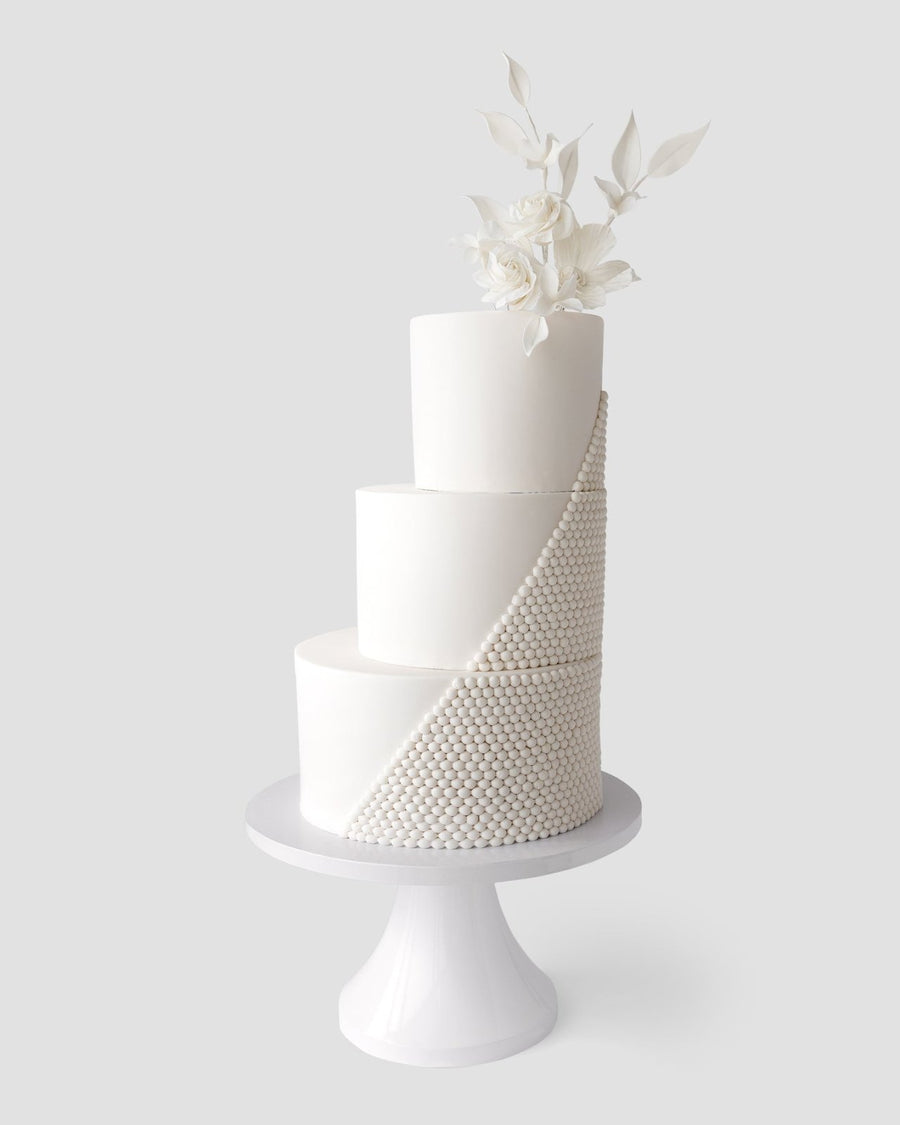 The Wedding Cakes Under $1,000 by Jenna Rae Cakes features a three-tiered white fondant cake with a diagonal cascade of pearl-like beads and white flowers with leaves, elegantly displayed on a white pedestal stand.