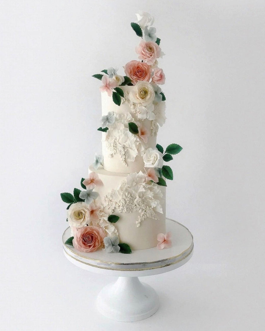The Jenna Rae Cakes Wedding Cakes Under $1,000 collection features elegant two-tiered white cakes, adorned with realistic pink, white, and peach sugar flowers with green leaves, beautifully displayed on a simple white cake stand.