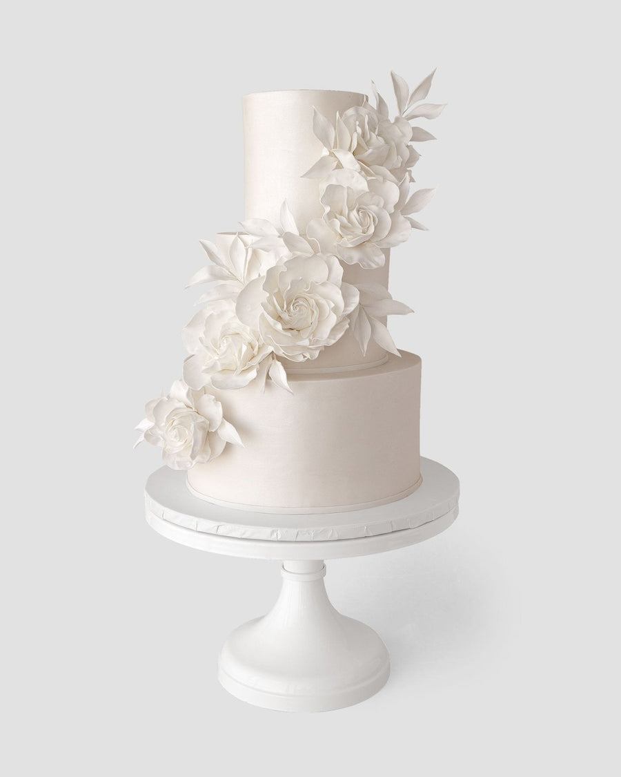 A three-tiered white cake by Jenna Rae Cakes, part of their Wedding Cakes Under $1,000 collection, features elegant white sugar flowers and is presented on a pedestal against a light gray background.