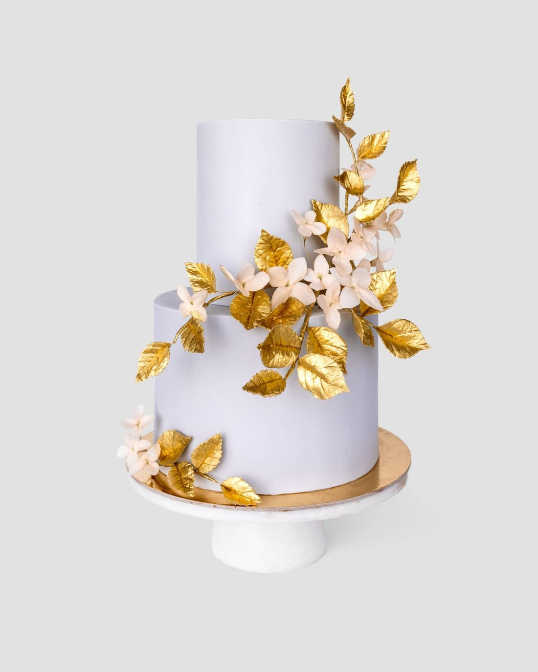 A two-tiered white cake with gold leaves and pale pink flowers from Jenna Rae Cakes Wedding Cakes Under $1,000 collection, shown on a white pedestal stand against a plain background.