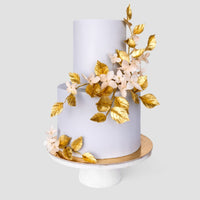A two-tiered white cake with gold leaves and pale pink flowers from Jenna Rae Cakes Wedding Cakes Under $1,000 collection, shown on a white pedestal stand against a plain background.