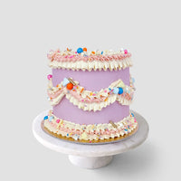 A pastel purple cake from Jenna Rae Cakes Wedding Cakes Under $200 collection, topped with pink and white frosting, sprinkles, and candies, sits on a marble stand against a light grey backdrop.