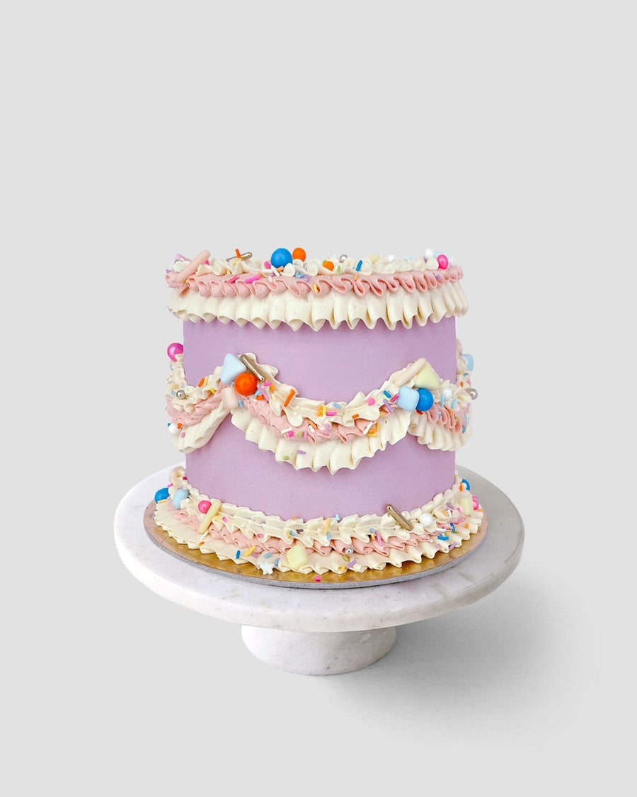 A pastel purple cake from Jenna Rae Cakes Wedding Cakes Under $200 collection, topped with pink and white frosting, sprinkles, and candies, sits on a marble stand against a light grey backdrop.