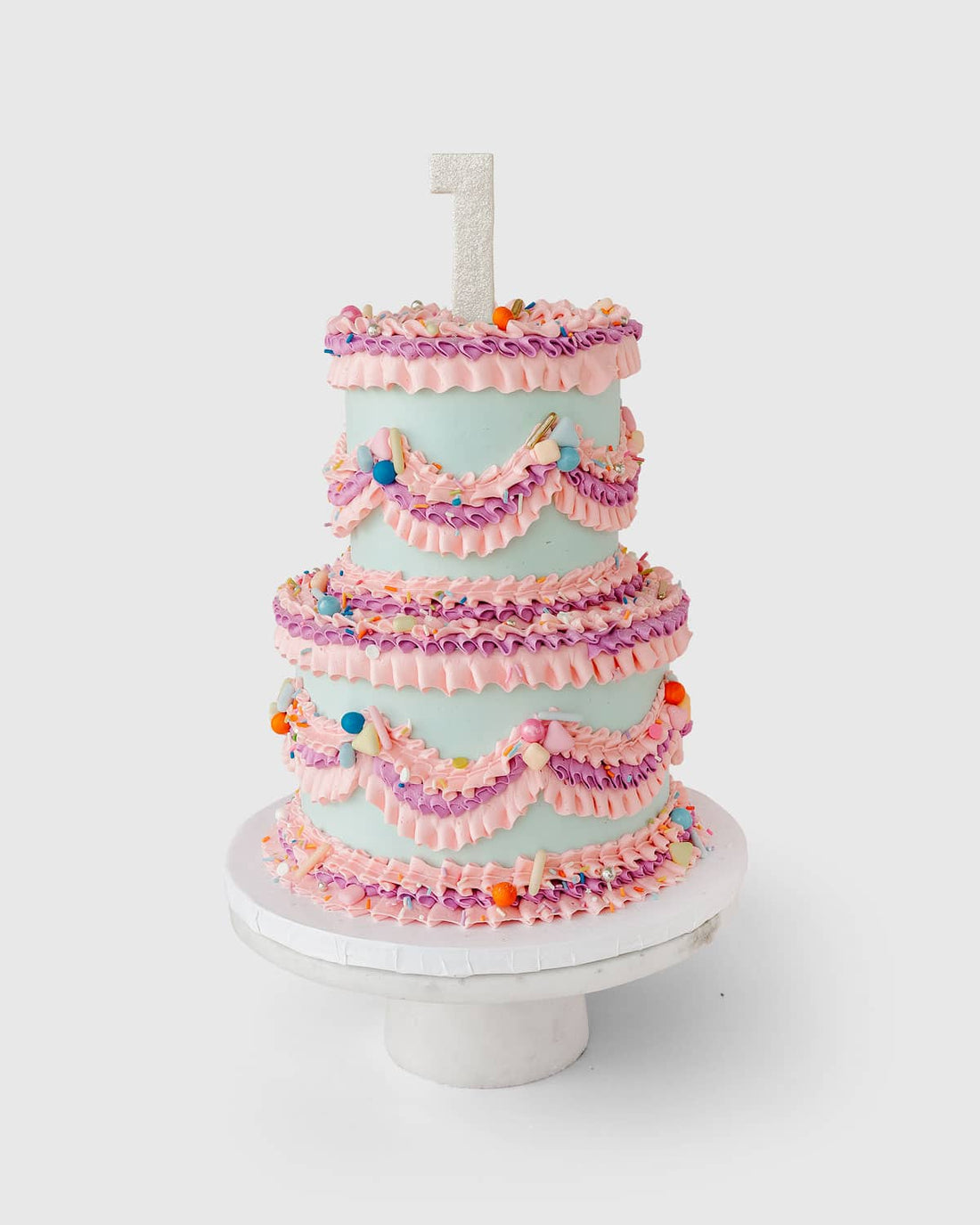 The Wedding Cakes Under $500 by Jenna Rae Cakes features three tiers, pink and purple icing swags, colorful sprinkles, a glittery number one candle topper, and comes displayed on a white cake stand.