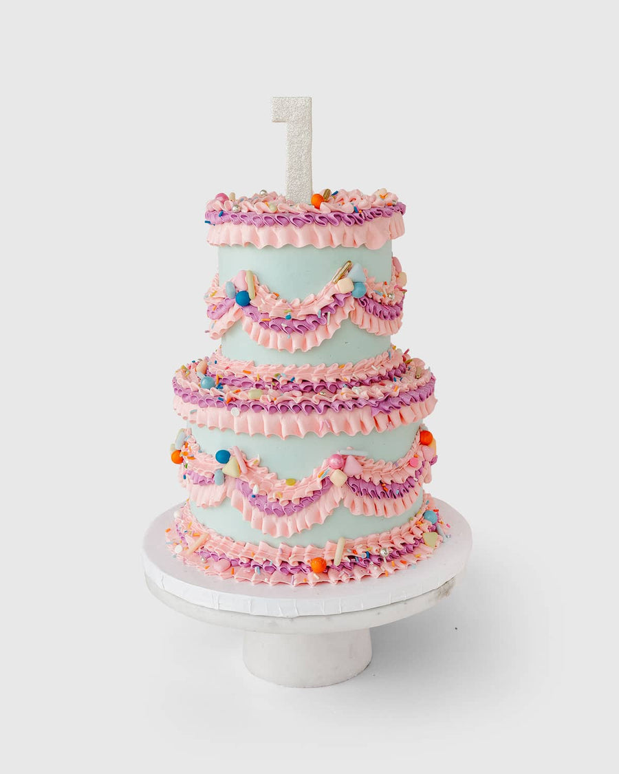 The Wedding Cakes Under $500 by Jenna Rae Cakes features three tiers, pink and purple icing swags, colorful sprinkles, a glittery number one candle topper, and comes displayed on a white cake stand.
